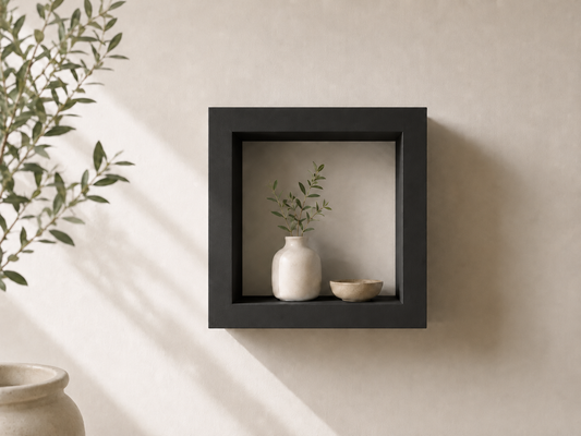 A square black floating wall shelf mounted on a textured beige wall, styled with a small white ceramic vase and a beige bowl containing green sprigs, with soft sunlight casting shadows.