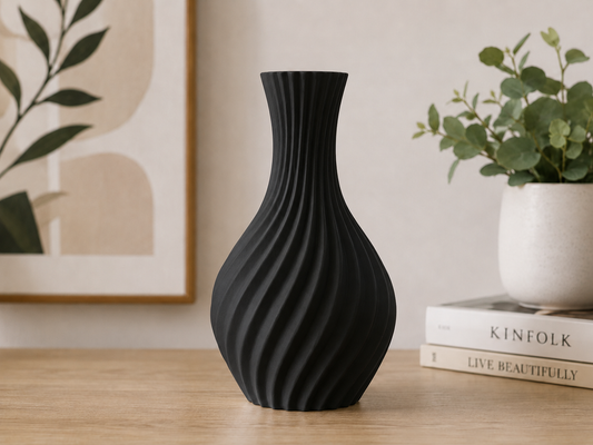 A matte black ribbed vase with a flared neck and bulbous base, standing on a light wood tabletop in a styled interior setting with a framed print, books, and a potted plant in the background.