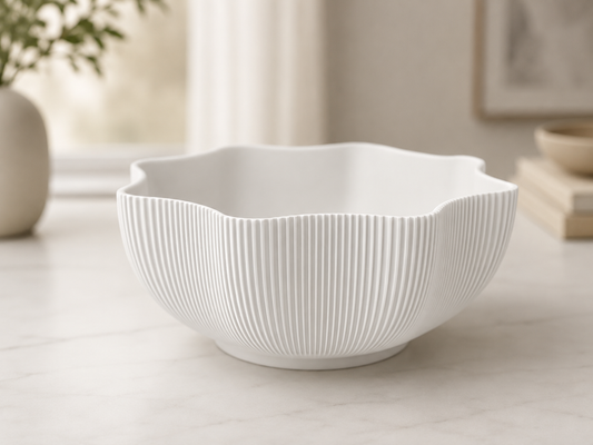 A white ceramic bowl with a scalloped rim and vertical fluted texture, shown on a light marble countertop in a softly lit kitchen setting.