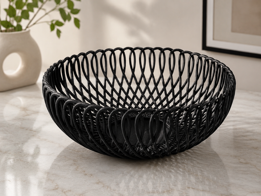 A black woven oval wire bowl with a basketweave pattern, displayed on a light marble surface with a blurred modern interior background.