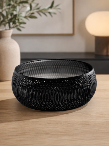A round black woven decorative bowl with a textured, open-weave pattern, displayed on a light wood tabletop in a modern, softly-lit interior setting.