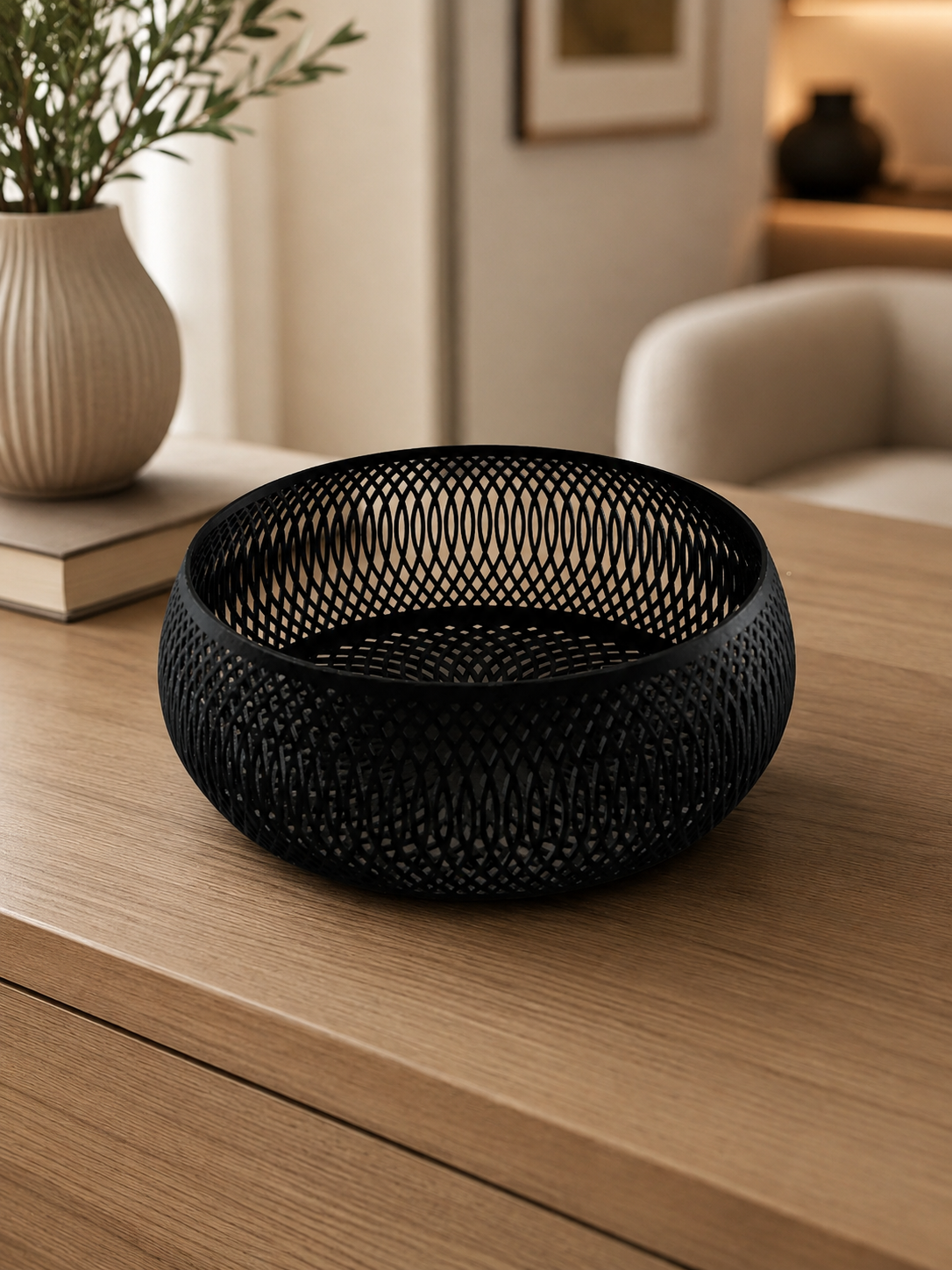 A black woven mesh bowl with a textured, open-weave pattern, displayed on a light wood table in a warm, modern living room setting.