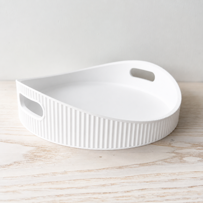 A white ceramic serving tray with a ribbed exterior and two oval cutout handles, shown on a light wood surface against a neutral light gray background.