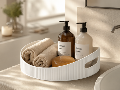 A white ribbed corner bathroom organizer tray holding two rolled beige towels, a round amber soap dish, and two Meraki toiletry bottles on a bathroom counter.
