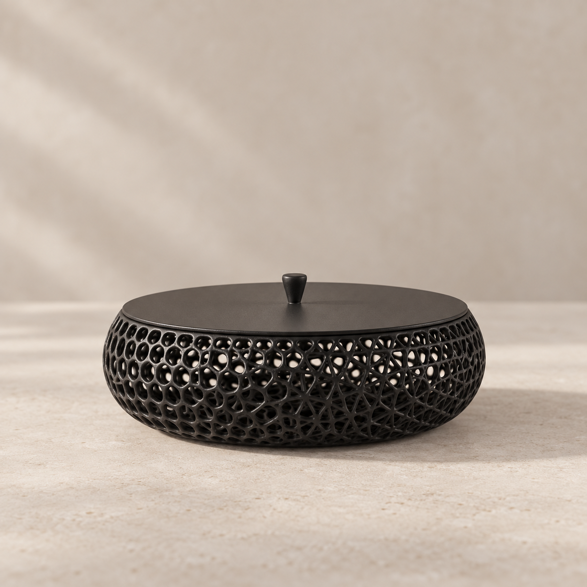 A round black metal decorative container with a perforated geometric lattice pattern and a matching lid, set against a neutral beige textured background.