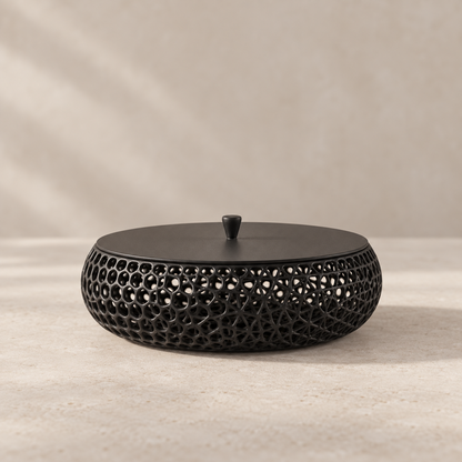 A round black metal decorative container with a perforated geometric lattice pattern and a matching lid, set against a neutral beige textured background.