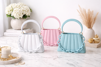 Three small handbags in white, pink, and blue on a marble surface with decorative items.