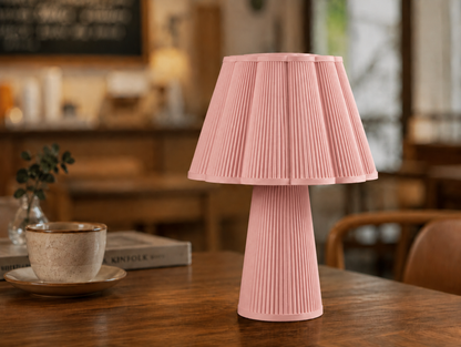 A pink pleated table lamp with a matching pleated base sits on a wooden table in a warm, softly blurred indoor setting with a cup and books nearby.