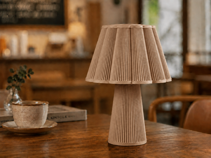 A pleated table lamp in a natural light wood finish, styled on a rustic wooden table next to a ceramic cup and saucer in a warm, softly blurred cafe interior.