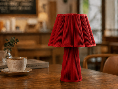 A red pleated table lamp with a matching pleated base sits on a wooden table in a warm, softly blurred indoor setting. A ceramic cup and saucer and a book are visible in the background.