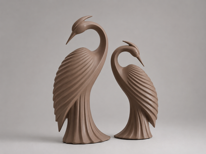 Two sculptural heron figurines in a matching warm taupe-brown glazed finish, standing side by side against a neutral light gray studio background.