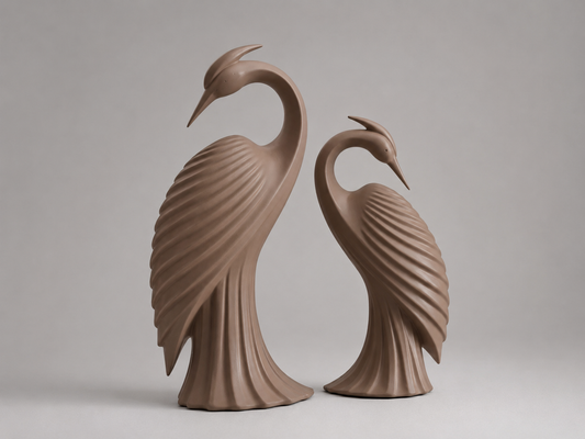 Two sculptural heron figurines in a matching warm taupe-brown glazed finish, standing side by side against a neutral light gray studio background.