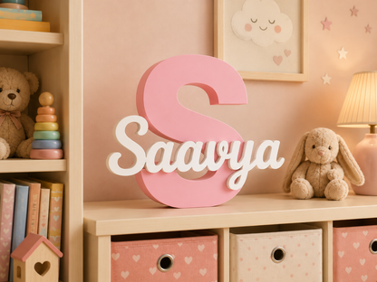 A large pink letter 'S' with the name 'Saavya' in white script, displayed on a wooden shelf in a cozy children's room with toys, a bunny plush, and pastel decor.