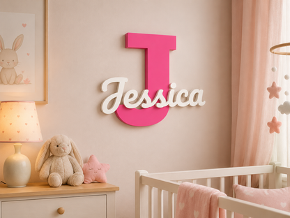 A pink and white personalized nursery wall decor piece featuring a large block letter 'J' and the name 'Jessica' in script, mounted on a beige wall above a white crib in a soft pink room.