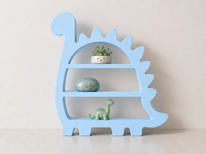 A light blue dinosaur-shaped wooden shelf with three tiers, styled with a small potted succulent, a blue-green stone, and a small green dinosaur figurine, set against a plain beige wall.