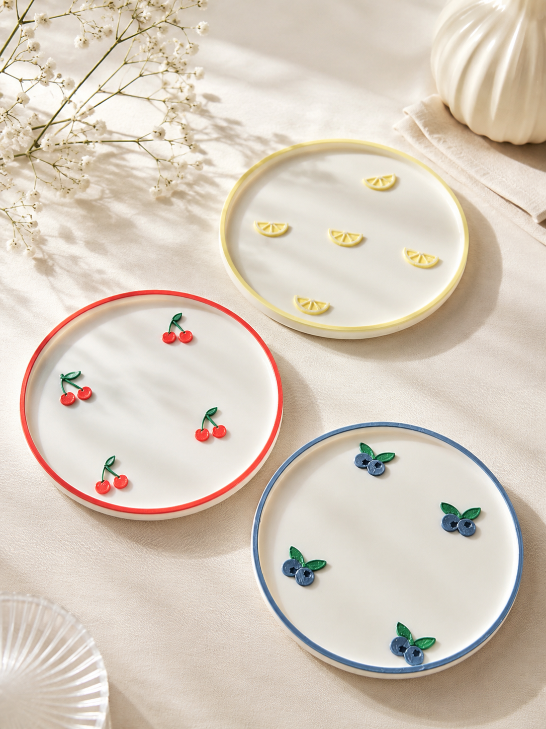 Three round white ceramic plates with colorful rims and small fruit motifs, arranged on a light beige fabric surface with soft shadows. One plate has red cherries, one has yellow slices, and one has blue berries.
