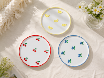Three round white decorative trays with cherry, lemon and blueberry motifs on a light beige fabric surface