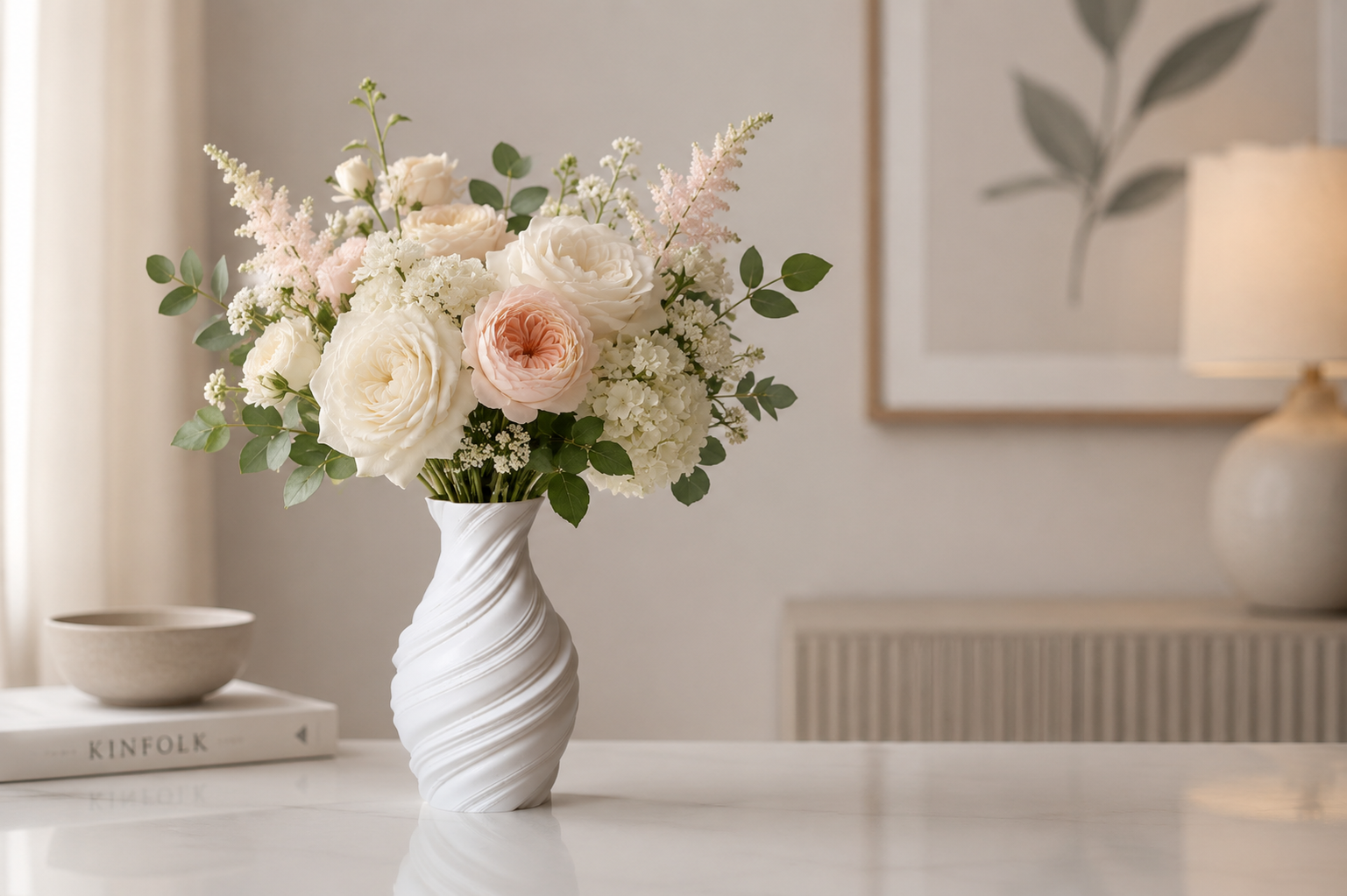 A white spiral-patterned ceramic vase holding a bouquet of white and blush pink roses with green foliage, placed on a white table in a softly lit, modern room.