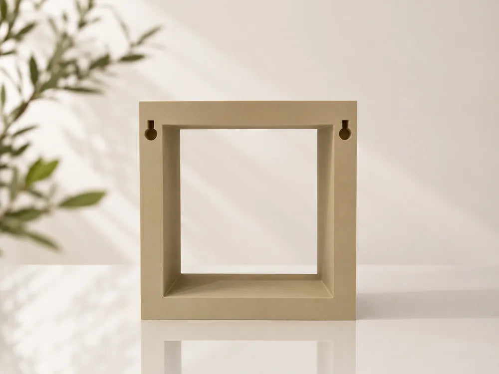 A square open-front shelf or frame in a warm beige tone, standing on a white surface against a light wall with soft shadows and blurred green leaves on the left.