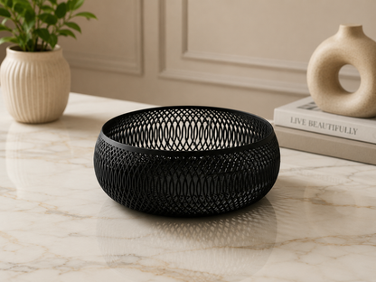 A black metal mesh bowl with a woven pattern, displayed on a light marble tabletop with a potted plant and decorative books in the background.