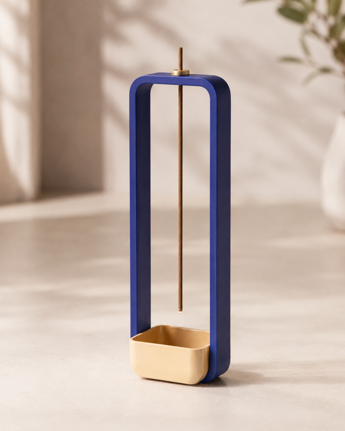 A modern blue and gold incense holder with a vertical frame, a single incense stick suspended in the center, and a small gold base, set against a softly lit neutral background.
