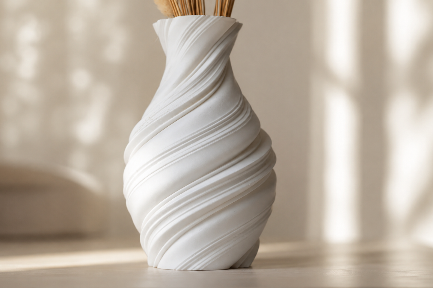 A white sculptural spiral vase with dried grass stems, set on a light wooden surface in a softly lit room with warm sunlight and shadows.