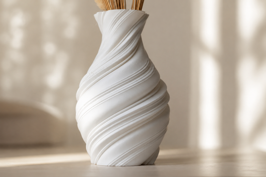 A white sculptural spiral vase with dried grass stems, set on a light wooden surface in a softly lit room with warm sunlight and shadows.