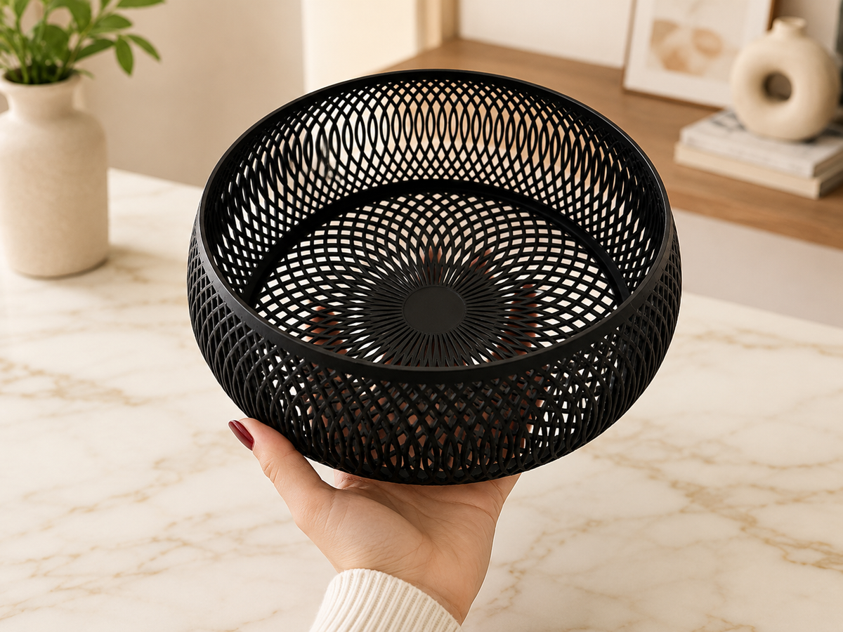 A hand holds a black metal mesh basket with a circular lattice pattern, set against a softly blurred indoor background with a marble surface.