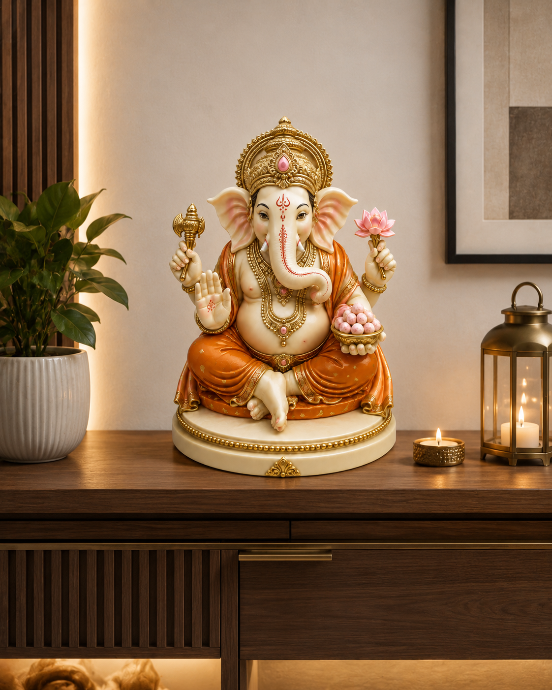 A decorative Ganesha statue seated on a wooden console table in a warmly lit interior. The statue is cream and white with gold detailing, orange drapery, and holds a pink lotus flower and a bowl of sweets.
