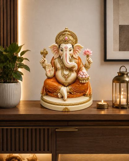A decorative Ganesha statue seated on a wooden console table in a warmly lit interior. The statue is cream and white with gold detailing, orange drapery, and holds a pink lotus flower and a bowl of sweets.