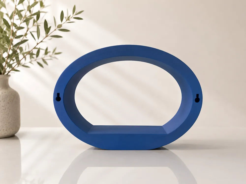 A blue oval-shaped wall-mounted shelf or decorative frame with two small keyhole cutouts on the sides, displayed on a white surface with a beige wall and a potted plant in the background.