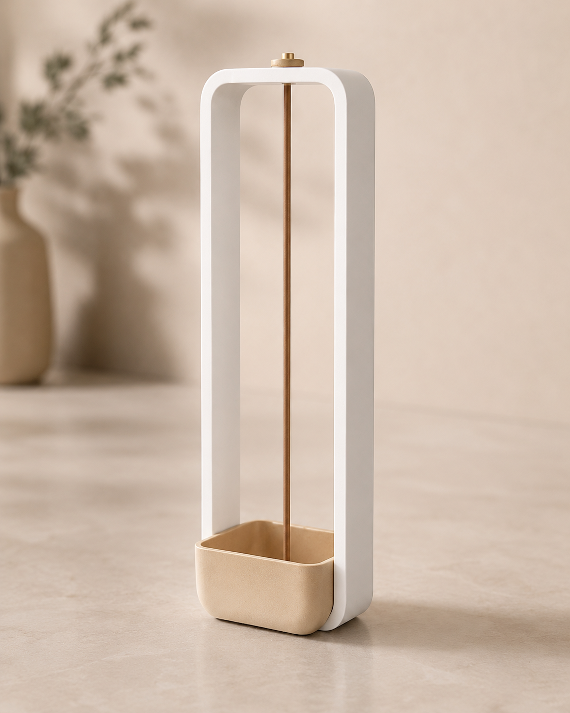 A minimalist incense burner with a white rectangular frame, a gold-toned incense stick, and a beige ceramic base, set against a warm beige background with a blurred vase in the distance.