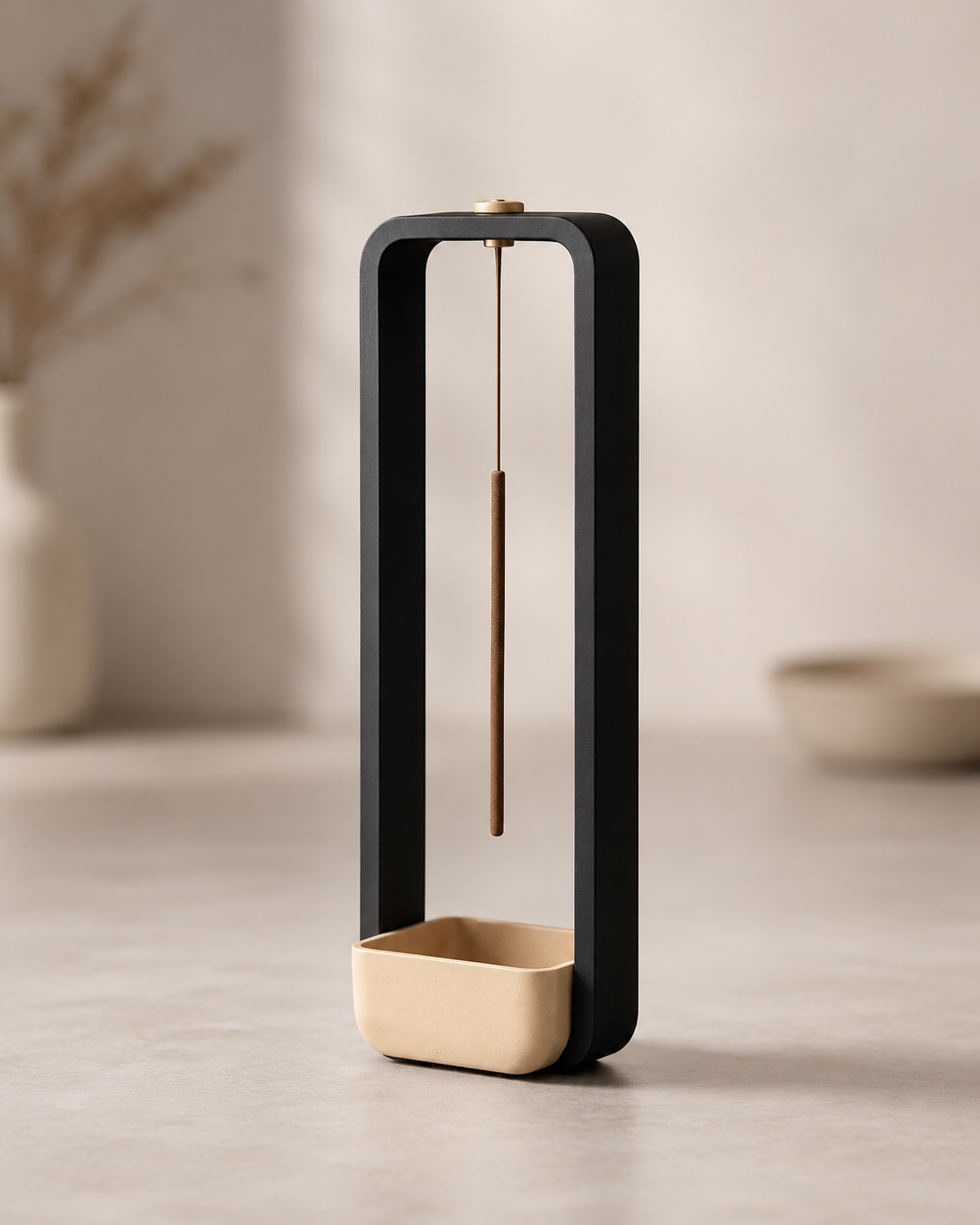 A modern minimalist tabletop clock with a matte black rectangular frame, a gold-toned hanging pendulum, and a gold-toned square base, set against a warm beige background with soft-focus decorative elements.