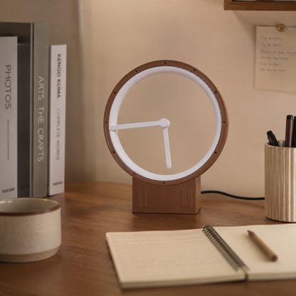 A modern round desk lamp with a warm brown frame and base, featuring a glowing white LED ring and white hands, styled on a wooden desk next to books, a notebook, and a cup.