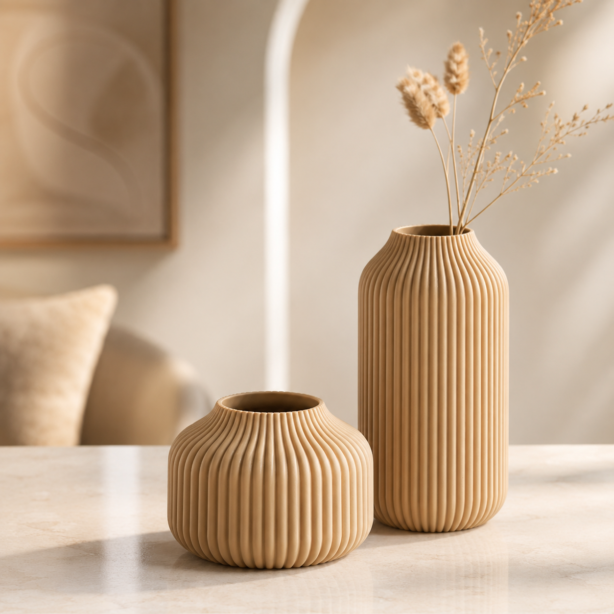 Two ribbed ceramic vases in warm beige tones, one small round vase and one taller narrow vase with dried stems, displayed on a light tabletop in a softly lit interior setting.