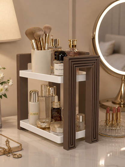 A two-tier cosmetic organizer with a dark brown frame and white shelves, styled on a vanity table with makeup brushes, skincare bottles, and perfume bottles. A round vanity mirror with a warm light is visible in the background.