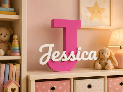 A large pink letter J with the name 'Jessica' in white script across it, displayed on a shelf in a pastel children's room with toys, a framed star print, and a lamp.