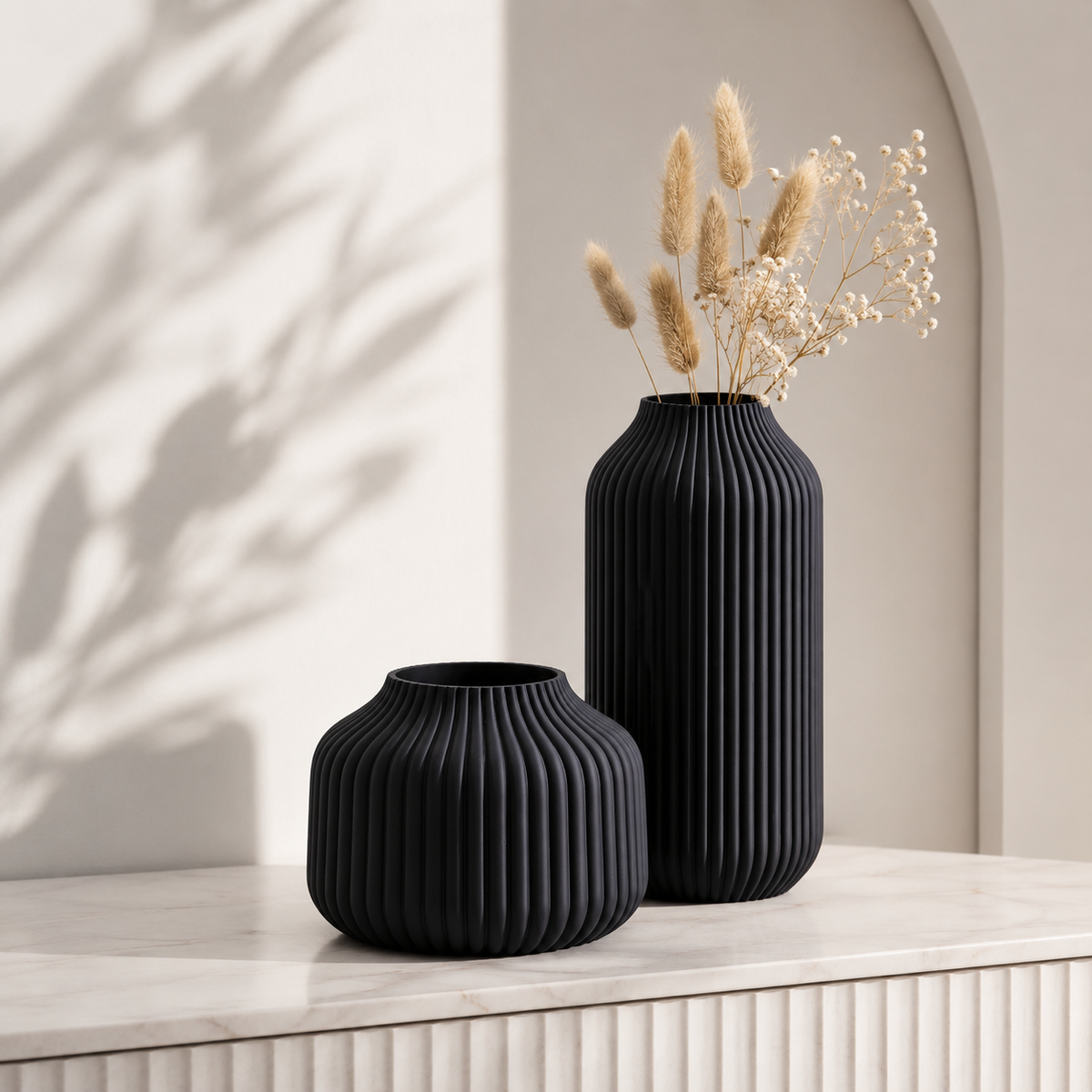 Two black ribbed ceramic vases on a light stone surface, with dried grass and baby's breath stems in the taller vase, against a warm beige wall with soft shadow patterns.