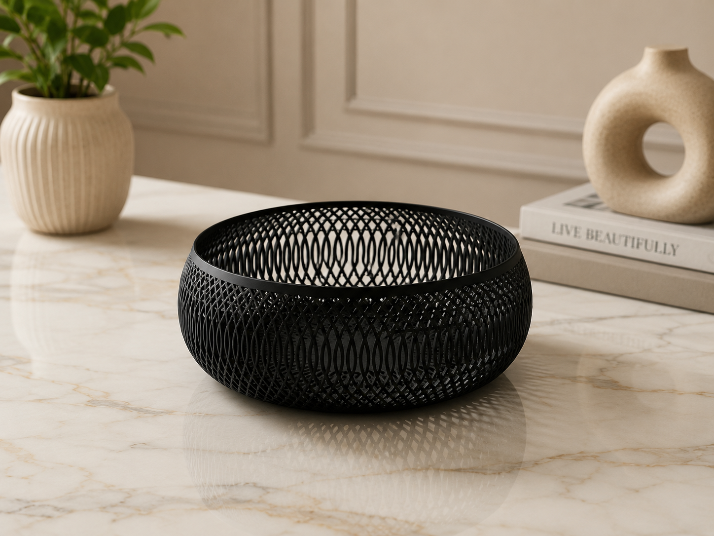 A black metal mesh bowl with a woven pattern, displayed on a light marble tabletop with a potted plant and decorative books in the background.