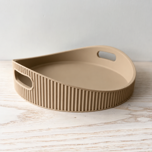 A beige silicone serving tray with a ribbed exterior and integrated handles, shown on a light wood surface against a neutral background.