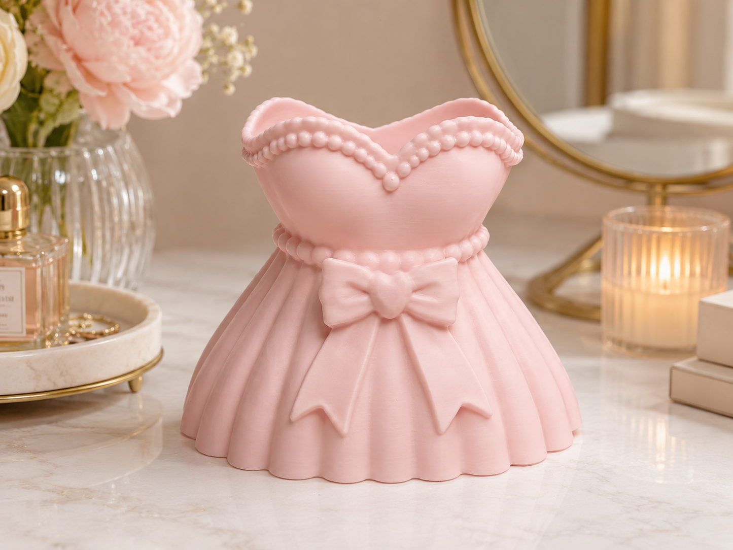 A pink dress-shaped decorative container or organizer sits on a marble surface in a softly styled interior setting with flowers, a candle, and a mirror in the background.