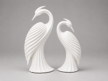A pair of white ceramic decorative bird sculptures shaped like stylized herons or cranes, standing side by side against a neutral gray studio background.