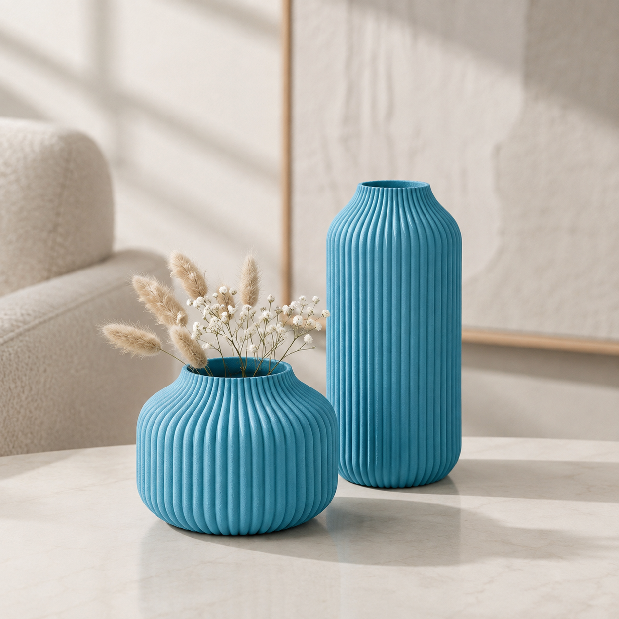 Two ribbed ceramic vases in bright turquoise blue, one short and wide with dried grasses inside and one tall and narrow, displayed on a light marble table in a softly lit interior setting.