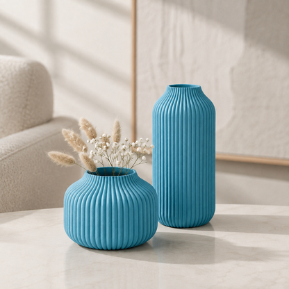 Two ribbed ceramic vases in bright turquoise blue, one short and wide with dried grasses inside and one tall and narrow, displayed on a light marble table in a softly lit interior setting.