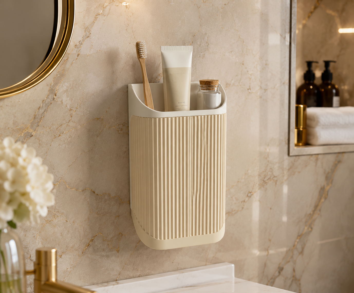 A wall-mounted bathroom organizer cup in a warm cream color, holding a wooden toothbrush and a toothpaste tube, mounted on a beige marble wall.