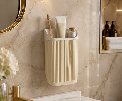 A wall-mounted bathroom organizer cup in a warm cream color, holding a wooden toothbrush and a toothpaste tube, mounted on a beige marble wall.