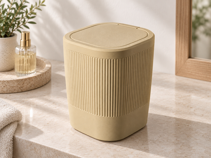 A beige ribbed cylindrical trash can with a matching lid sits on a light stone countertop in a bright bathroom setting, with a plant, folded towel, and small bottle in the background.