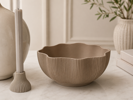 A beige ribbed ceramic bowl with a scalloped rim sits on a light marble surface in a styled interior setting with neutral decor.