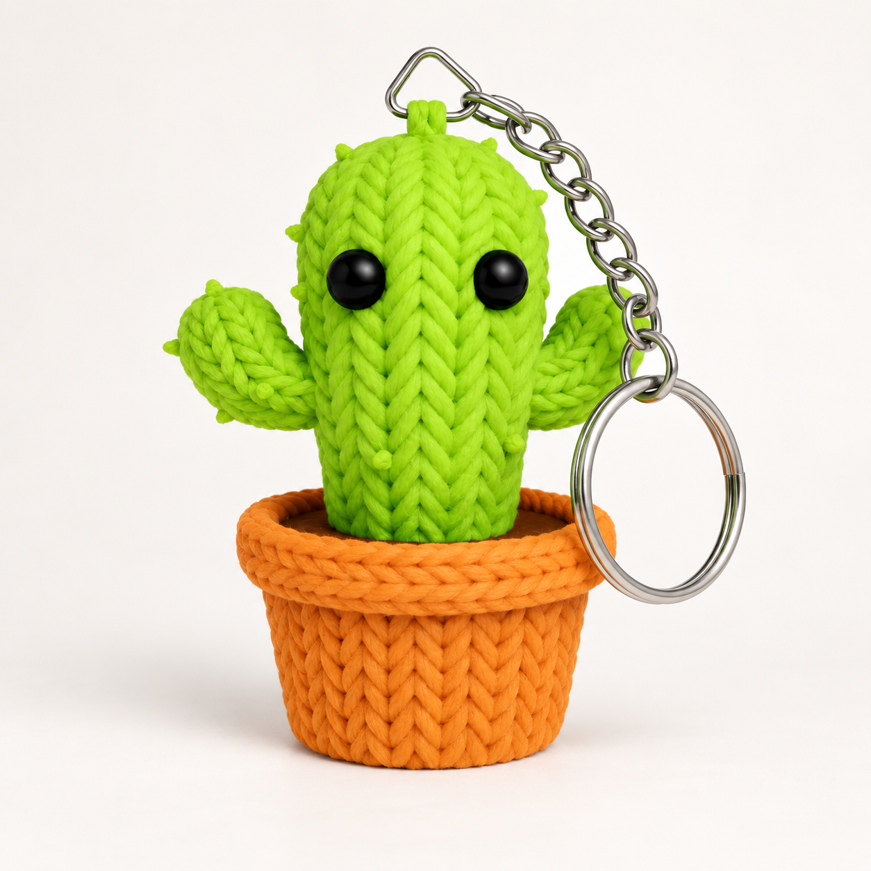 A knitted cactus keychain with a bright green cactus body, black eyes, and a silver metal keyring, sitting in an orange knitted pot on a clean white background.