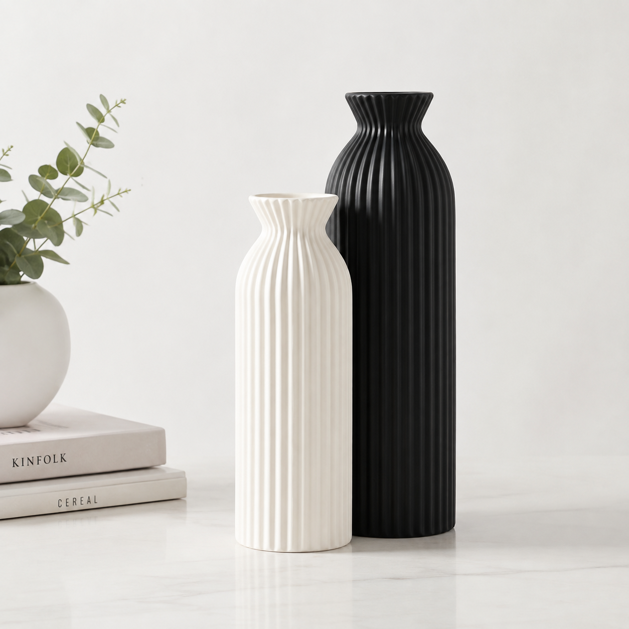 Two ribbed ceramic vases, one black and one off-white, standing on a light marble surface next to a small white planter with greenery and stacked books.